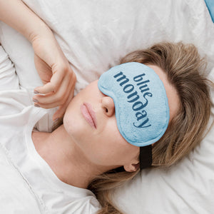 Person wearing a blue sleep mask with 'blue monday' text, lying on a white pillow.
