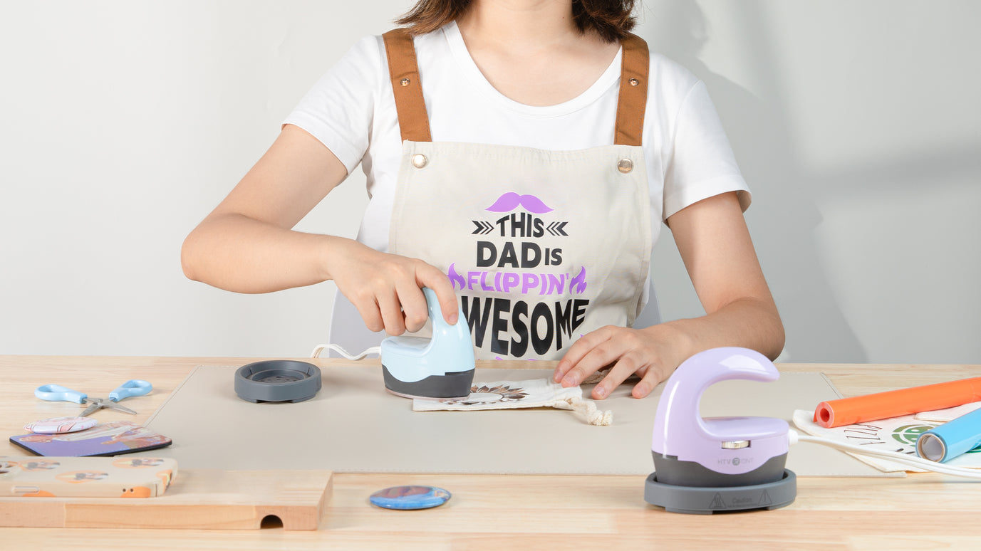 Person wearing a white apron with text, working at a table with H1 Mini Heat Press and materials.