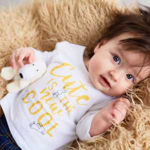 Baby wearing a white shirt with yellow text, lying on a fluffy beige surface.