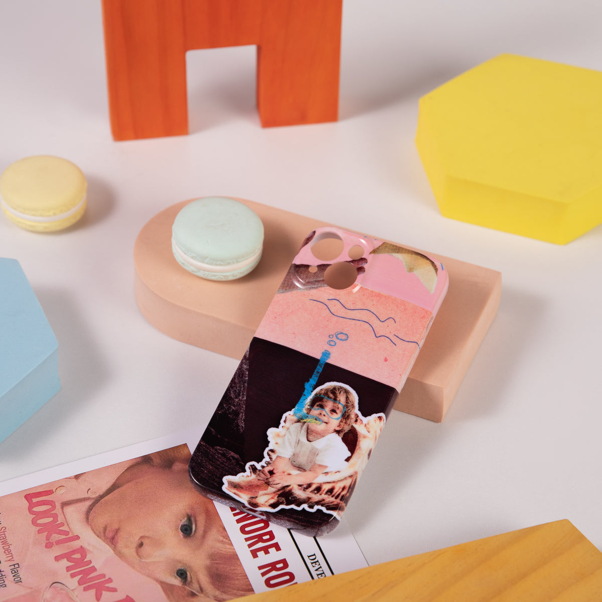Phone case with a design of a child holding a toy, placed on a colorful surface with macarons and a magazine.