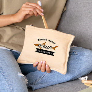 Person holding a beige pouch with 'Wonder World Ocean Logbook' text, sitting on a gray couch.