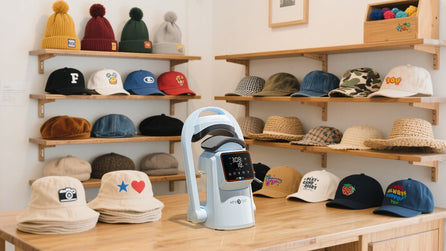 A light blue A300 Hat Press sits in the middle of the table, with various DIY hats hanging behind it.