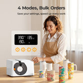 Woman in an apron using a white appliance with digital display, surrounded by colorful mugs.
