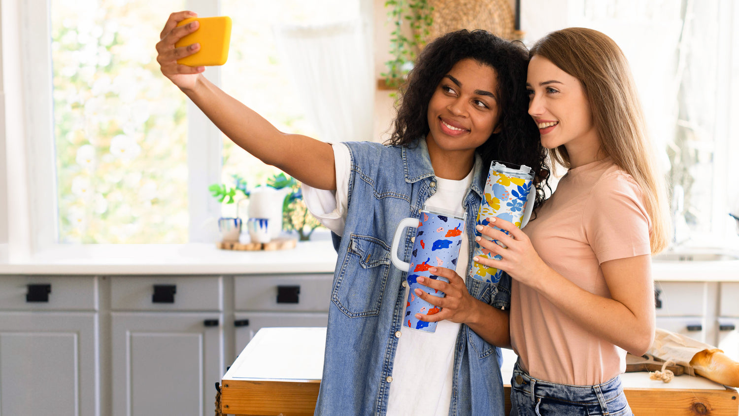 Two women taking a selfie in a kitchen holding colorful mugs.