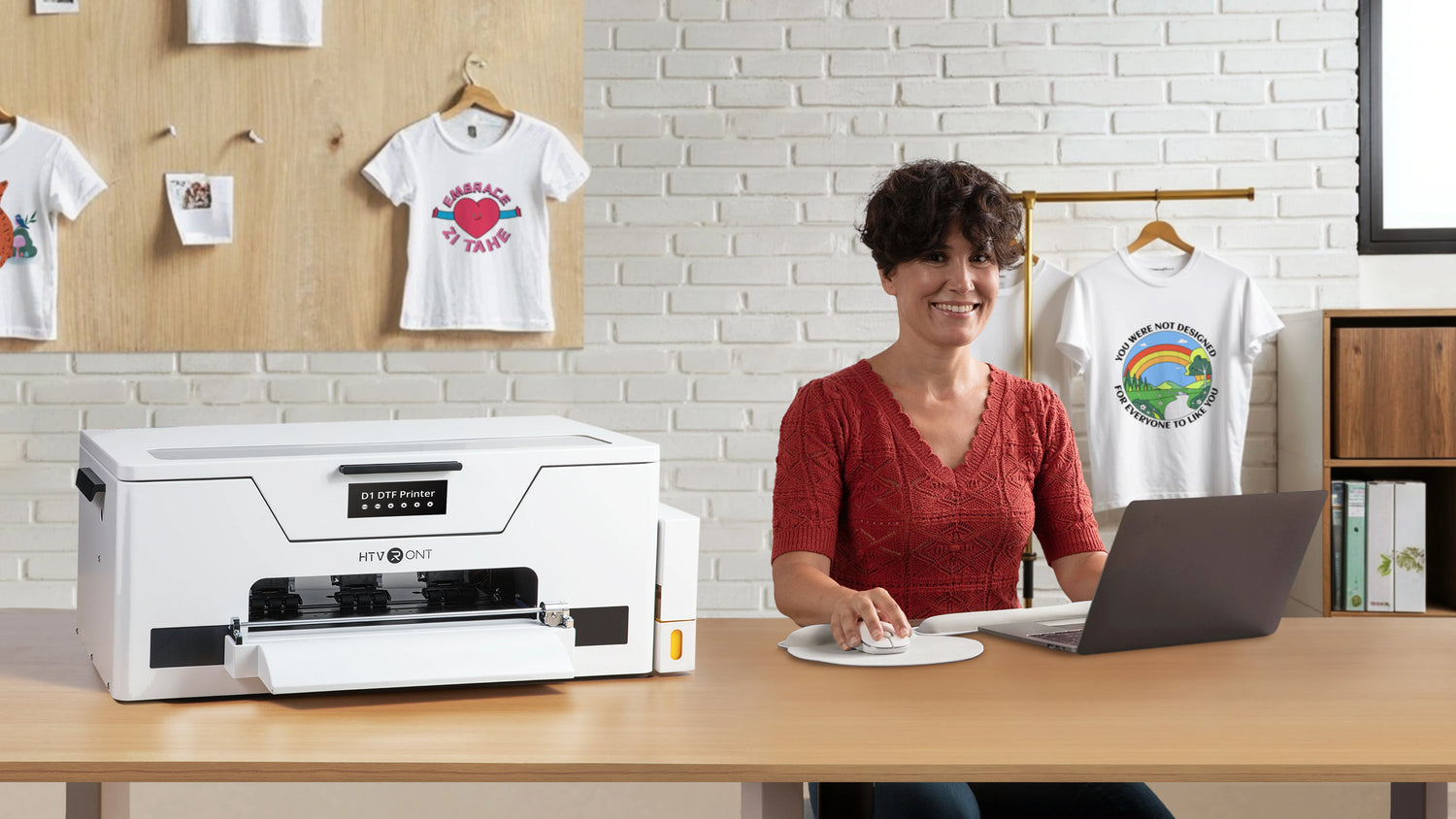 Woman using a laptop next to a white D1 DTF printer with t-shirts in the background.